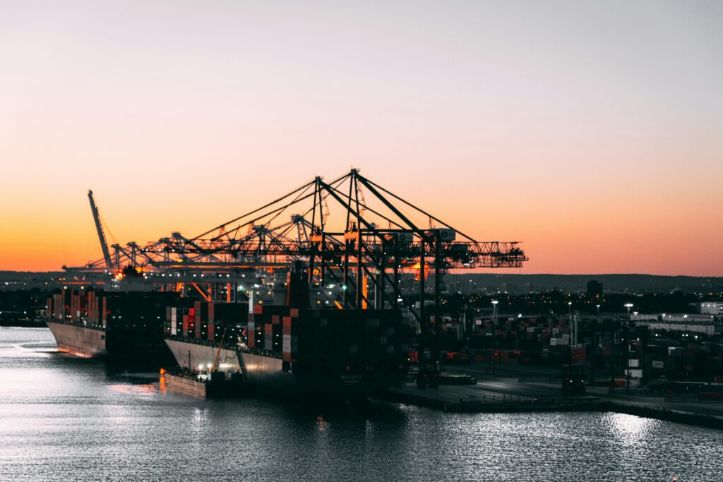 Home A serene view of cargo ships and cranes at a harbor during sunset, highlighting maritime industry.